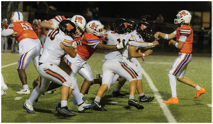 A group of people in football uniforms are engaged in a play on a lit field at night with one person throwing a football. Description generated by AI