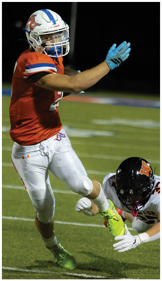A person wearing a red football jersey and white pants is running on a field while another person in a dark uniform reaches out toward them. Description generated by AI