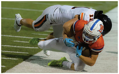 Two people in football uniforms are engaged in a tackle near the sideline on a grassy field during a game. Description generated by AI