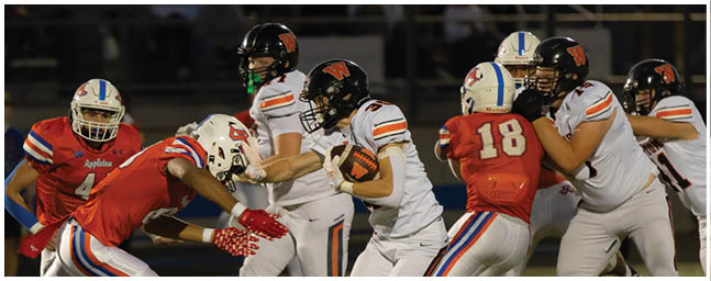 People in football uniforms are actively engaged in a play on a field under stadium lights. Description generated by AI