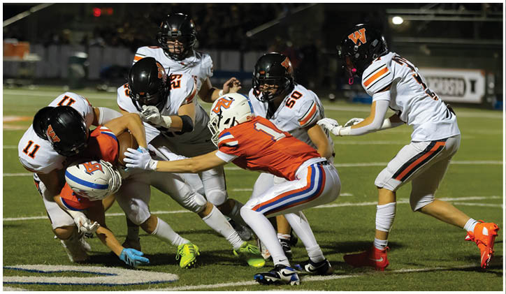 Several people in football uniforms are engaged in a tackle on a grassy field near yard line markings. Description generated by AI