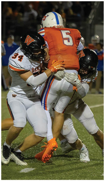 Three people in football uniforms are engaged in a tackle on a grassy field during a game. Description generated by AI