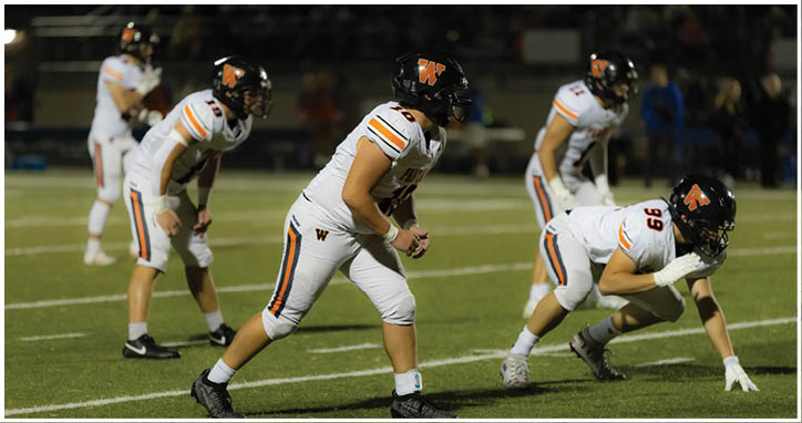 Four people in white football uniforms and black helmets are positioned on a football field, preparing for a play during a game. Description generated by AI
