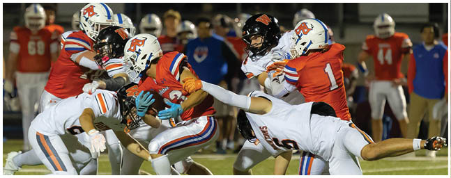 Players in red and white football uniforms are engaged in a tackle during a nighttime game on a field. Description generated by AI