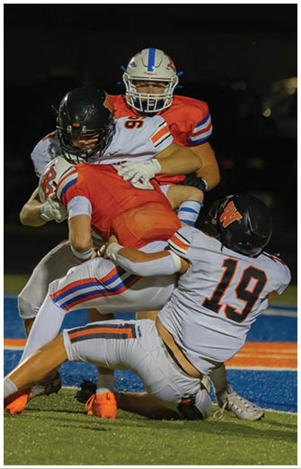 Four people in football uniforms are engaged in a tackle on a field with blue and orange markings during a nighttime game. Description generated by AI