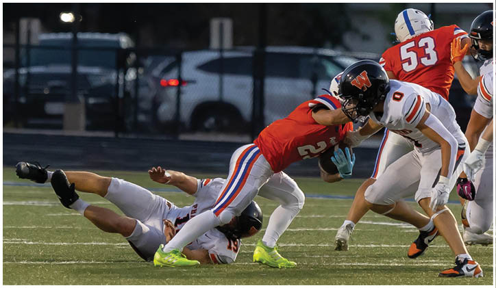 A person in a red sports uniform is running with a football while two people in white uniforms attempt to tackle them on a football field. Description generated by AI