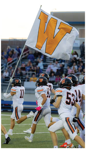 A group of football players in white uniforms with black helmets and orange accents are running on a field with a large flag displaying a bold orange “W" in the foreground and a crowd of spectators in the background. Description generated by AI
