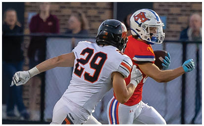 Two people in football uniforms are running on a field with one person attempting to catch or tackle the other who is holding a football. Description generated by AI