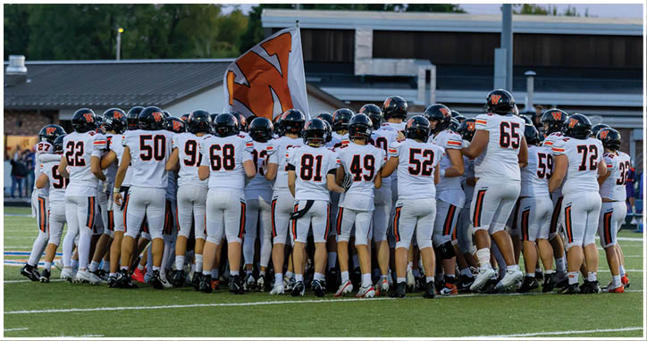 A group of adults wearing white football uniforms with black helmets are huddled together on a football field with a flag in the background. Description generated by AI