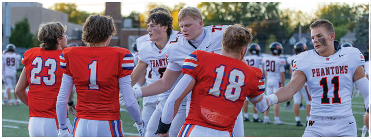 Five people wearing football uniforms are standing on a field, with some shaking hands and others facing each other. Description generated by AI
