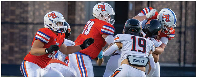 Four people wearing football helmets and uniforms are engaged in a play on a field, with two in red jerseys and two in white jerseys. Description generated by AI