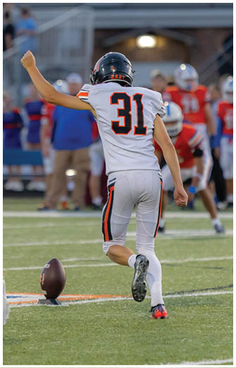 A person wearing white athletic shoes is about to kick a football placed on a tee on a grassy field with blurred people and benches in the background. Description generated by AI
