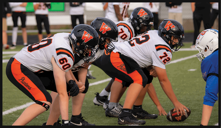 Several people in football uniforms are lined up on a field in a ready stance facing opponents in blue uniforms during a game. Description generated by AI