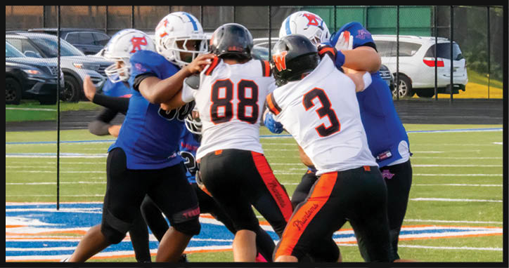 Several people wearing football uniforms are engaged in a play on a green field with cars visible in the background. Description generated by AI