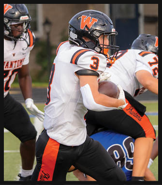 People wearing football helmets and uniforms are engaged in a game, with one person holding a football and others reaching out. Description generated by AI