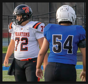 Two people wearing football uniforms stand on a field, one in a white jersey with the number 72 and the word “Phantoms" and the other in a blue jersey with the number 54. Description generated by AI