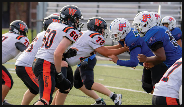 People in football uniforms are engaged in a play on a grassy field with some players in white and orange and others in blue jerseys. Description generated by AI