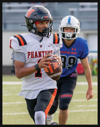 A person wearing a white football uniform with “Phantoms" on the front is holding a football and running on a field while another person in a blue uniform follows behind. Description generated by AI
