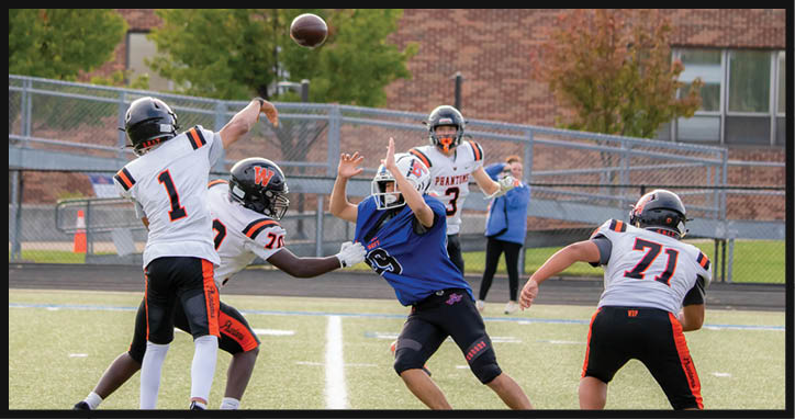 Four people in football uniforms are actively engaged in a play on a field with one person in a blue jersey reaching out while the others in white jerseys prepare to block or pass. Description generated by AI