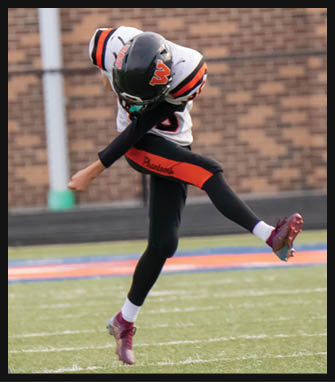 A person wearing a football uniform is kicking a football on a grassy field with a brick wall in the background. Description generated by AI