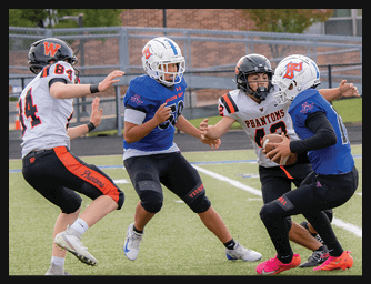 Four people wearing football helmets and uniforms are actively engaged in a play on a football field, with one person holding the ball while others attempt to block or tackle. Description generated by AI