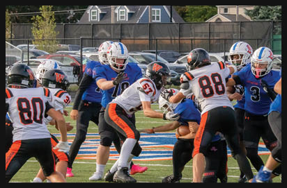 People wearing football uniforms are engaged in a play on a grassy field with a background of parked cars and houses. Description generated by AI
