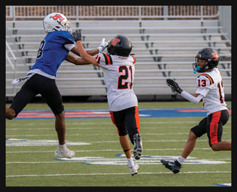 Three children in football uniforms are actively reaching for a football on a field with empty bleachers in the background. Description generated by AI