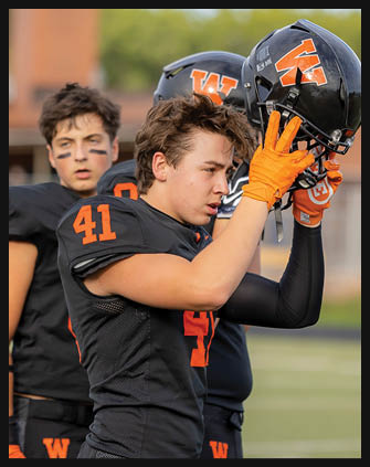 Three adults in black and orange football uniforms stand on a field, with one holding a helmet and wearing bright orange gloves. Description generated by AI