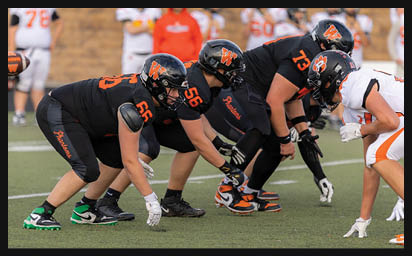 People in football uniforms are lined up on a field facing each other in a ready stance before a play. Description generated by AI