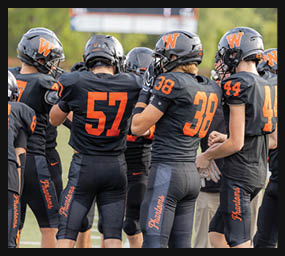 A group of people wearing black football uniforms with orange numbers and helmets are standing together on a field. Description generated by AI