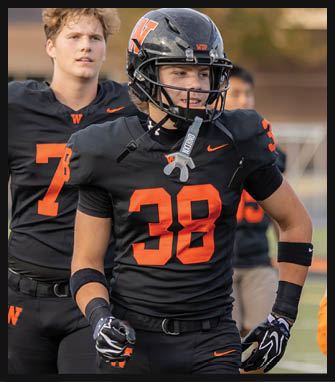 Two people wearing black and orange football uniforms stand on a field with one wearing a helmet and gloves while the other stands behind. Description generated by AI
