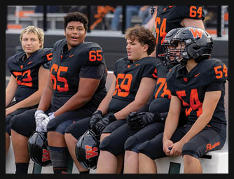A group of six people in black and orange football uniforms are sitting and standing on a bench at the side of a football field with a crowd in the background. Description generated by AI