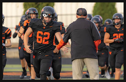 A group of people in black and orange football uniforms are running on a field while one person in a black jacket and cap reaches out to them. Description generated by AI