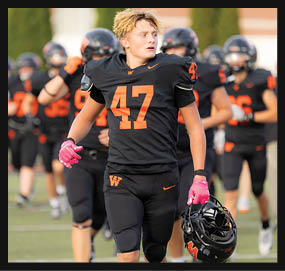 A person wearing a black football uniform with the number 47 and pink gloves is holding a helmet and walking on a field with other players and a coach in the background. Description generated by AI