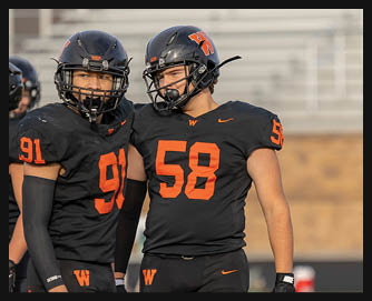 Two adults wearing black football uniforms with orange numbers and helmets stand closely together on a field. Description generated by AI