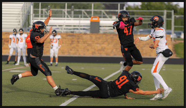 Four people in football uniforms are actively engaged in a play on a field with one person throwing a football while others are reaching or diving to catch or block it. Description generated by AI