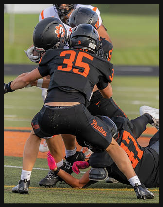 Several people wearing black and orange football uniforms are engaged in a play on a football field, with one person on the ground and others surrounding them. Description generated by AI