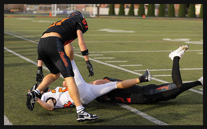 Three people in football uniforms are on a field with two lying on the ground and one standing over them during a game. Description generated by AI