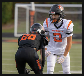Two people in football uniforms are facing each other on a field, one in white and orange and the other in black and orange, appearing ready for a play. Description generated by AI