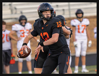 An adult wearing a black football uniform with the number 12 is preparing to throw a football on a field while other players and a referee stand in the background. Description generated by AI