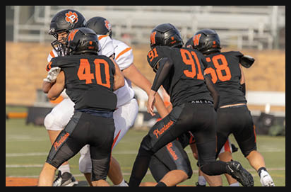 People wearing black and orange football uniforms are engaged in a play on a football field with others in white uniforms. Description generated by AI