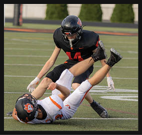 Two people in football uniforms are engaged in a play on a field, with one person on the ground and the other reaching toward them. Description generated by AI