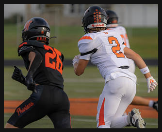 Two people in football uniforms are running on a field with one wearing black and orange and the other wearing white and orange. Description generated by AI