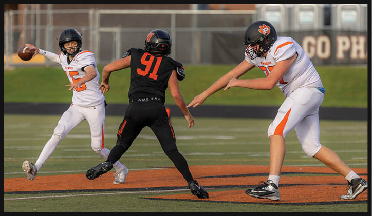Three people in football uniforms are on a field with one person preparing to throw a football while another person in black attempts to block and a third person in white braces for action. Description generated by AI