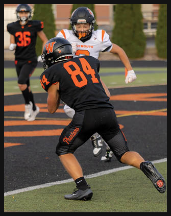 Two people in football uniforms are on a field with one person in black and orange running while the other in white and orange approaches from behind. Description generated by AI