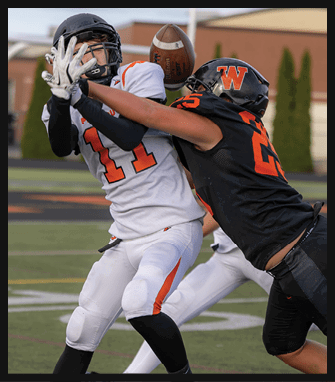 Two people in football uniforms are engaged in a physical play on a field, with one person in a black uniform tackling another in a white uniform holding a football. Description generated by AI