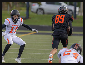 A person wearing a black football uniform with the number 89 is running on a field while two people in white and orange uniforms are attempting to catch or block them. Description generated by AI