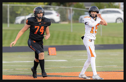 Two people wearing football uniforms and helmets are on a field, with one preparing to throw a football and the other running nearby. Description generated by AI