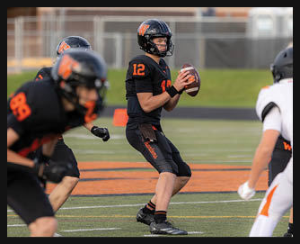 A person wearing a black football uniform with the number 12 is holding a football and preparing to throw it on a football field while other players in black and white uniforms are nearby. Description generated by AI