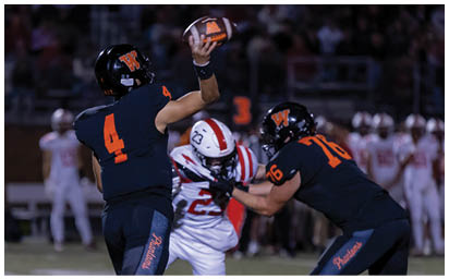 Two people in dark sports uniforms are blocking a person in a white sports uniform who is running with a football on a field during a game. Description generated by AI